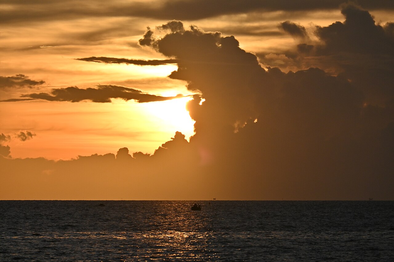 海面夕陽被積雨雲層遮蔽,金橙色光芒穿透烏雲,颱風季海景