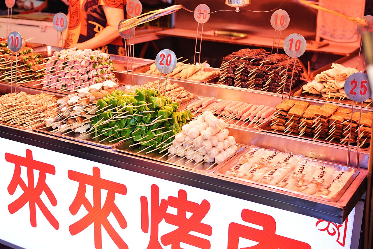 花蓮東大門夜市的烤肉攤特寫，不鏽鋼餐檯上一排排插著竹籤的肉串、豆皮、貢丸與時蔬，每盤插著圓形號碼價目牌，攤位下方是紅底白字的「來來烤肉」招牌。