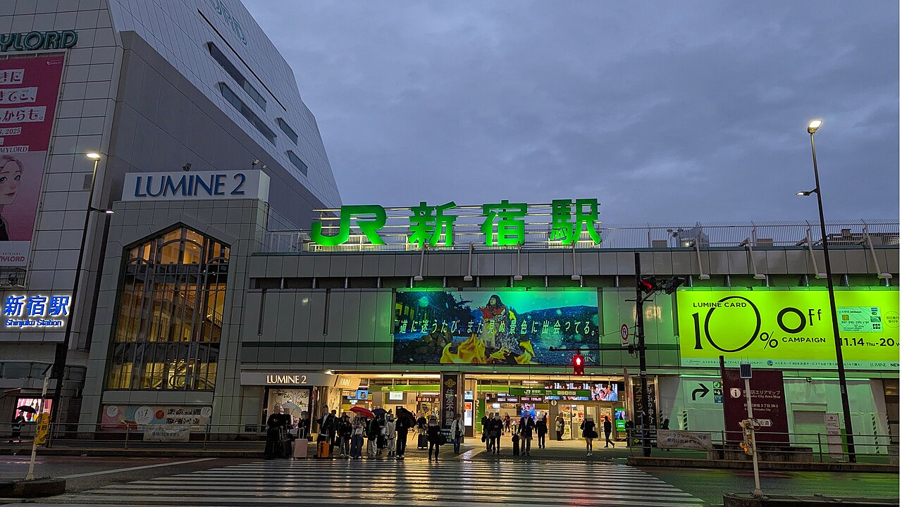 傍晚雨後的 JR 新宿站南口,車站上方是綠色的「JR新宿駅」大字,左邊是 LUMINE 2,右邊整面螢光綠的 LUMINE CARD 廣告牆,行人拖著行李走過濕漉漉的斑馬線。