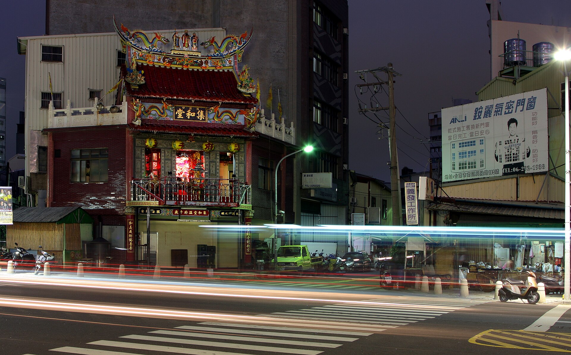 夜裡的台南街景，一棟紅色木構的老式廟宇建築亮著燈，夾在左右兩側的現代大樓中間，前方馬路上被車燈拖出一整條白色光軌，路邊停著機車，斑馬線橫過畫面。