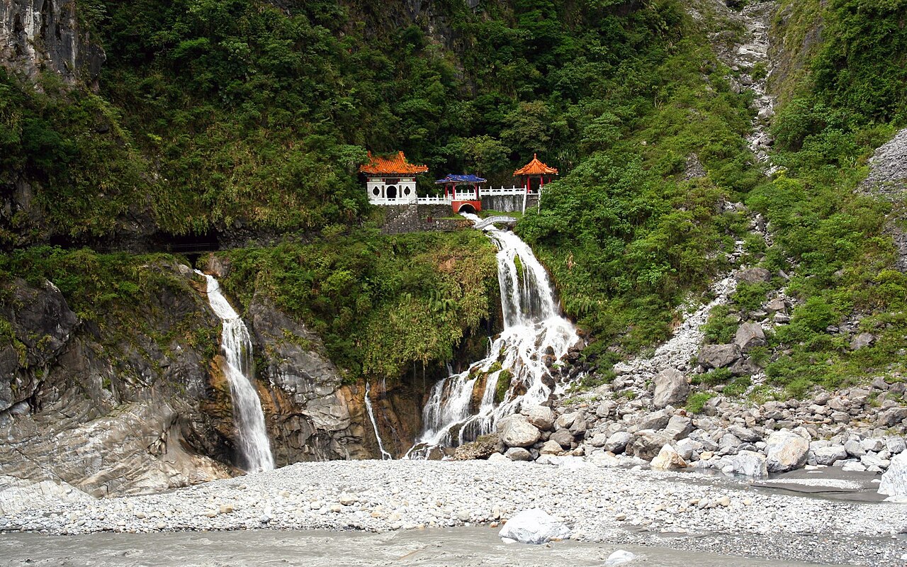 太魯閣長春祠，白牆橘瓦的中式廟宇建築群嵌在整片翠綠山壁的高處，下方兩道瀑布順著岩壁傾瀉而下，匯入佈滿白色礫石的溪谷。