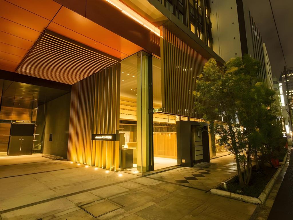 夜間的飯店入口,天花板是木格柵搭暖黃燈,整面玻璃門,門口地面鋪磁磚、旁邊種了小灌木。