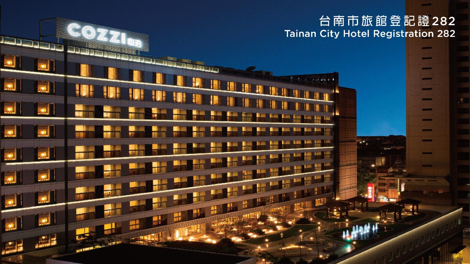 夜裡的飯店外觀，長條形建築的一整排房間都亮著暖黃色的燈，屋頂立著白色 COZZI 大字，樓下是亮著燈的街區。