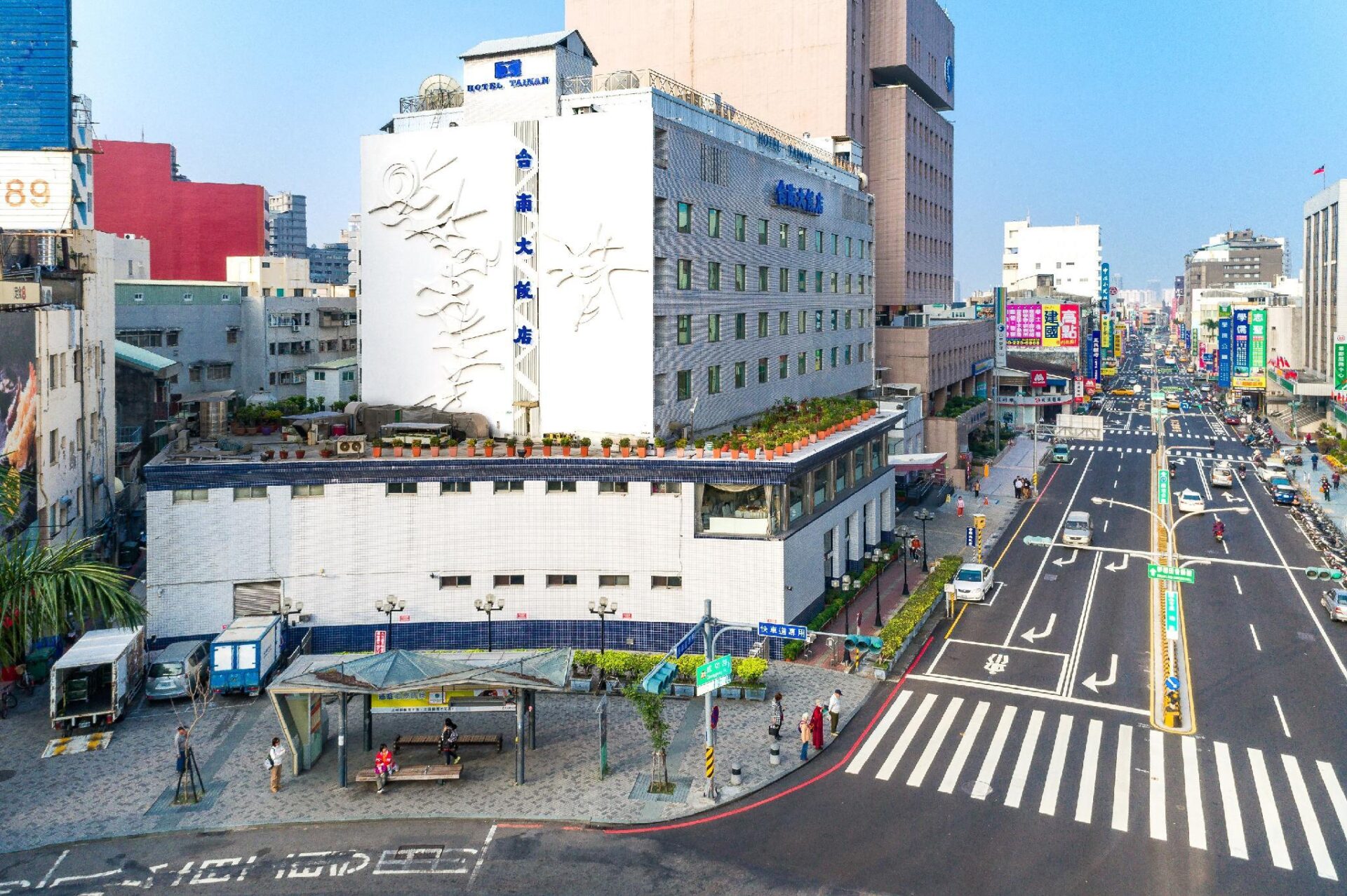 從高處往下拍的成功路口，白色建築側面掛著「台南大飯店」直式招牌，樓下是大馬路、斑馬線、公車候車亭與往來車流，遠方是密集的市區街景。