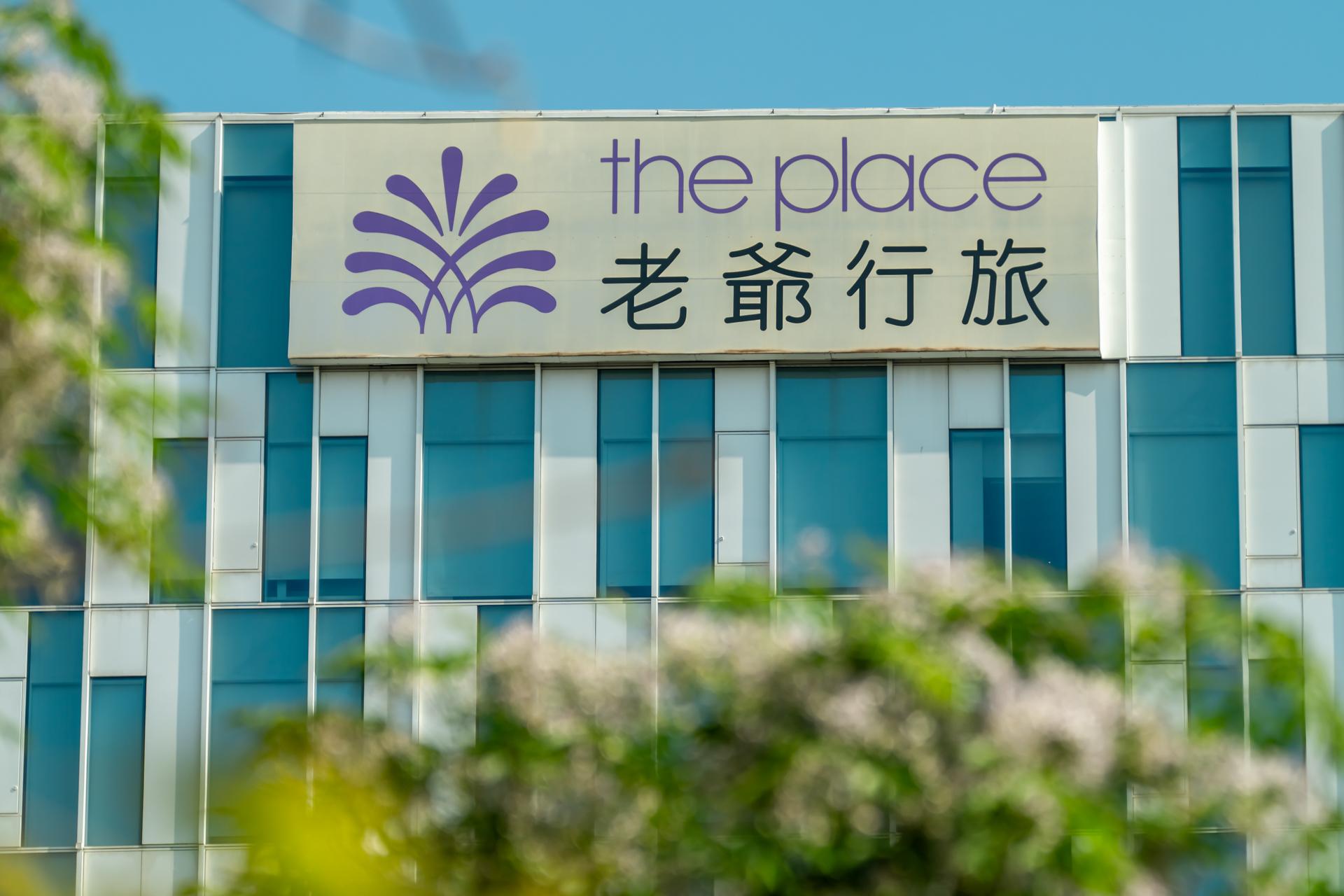 建築的藍綠色玻璃帷幕外牆上掛著一面白底招牌，寫著 the place 老爺行旅與紫色圖騰，前景是一叢開著白花的枝葉。