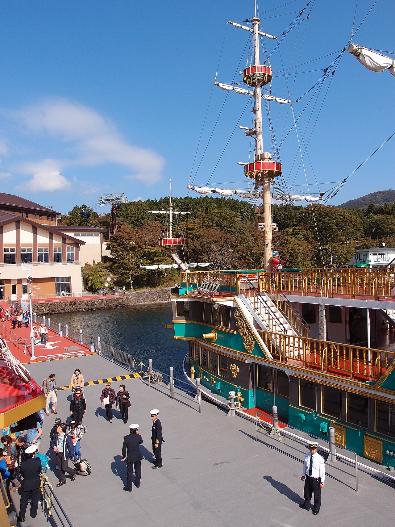 停靠中的箱根海賊船，制服船員在碼頭引導旅客上下船；畫面左後方的山坡上可以看到空中纜車的纜線與車廂。這一段（桃源台↔元箱根／箱根町）的成人單程票，2025/10/1 起從 1,200 円調為 1,700 円（示意圖，非本人拍攝）